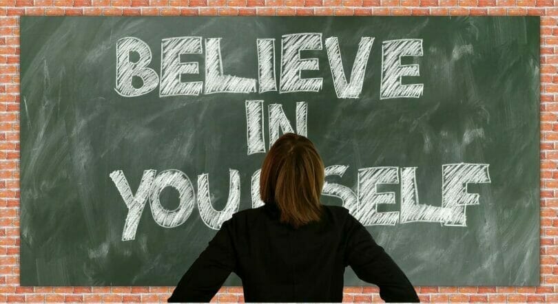 Person facing a chalkboard with "BELIEVE IN YOURSELF" written in large white letters on a green background against a brick wall.