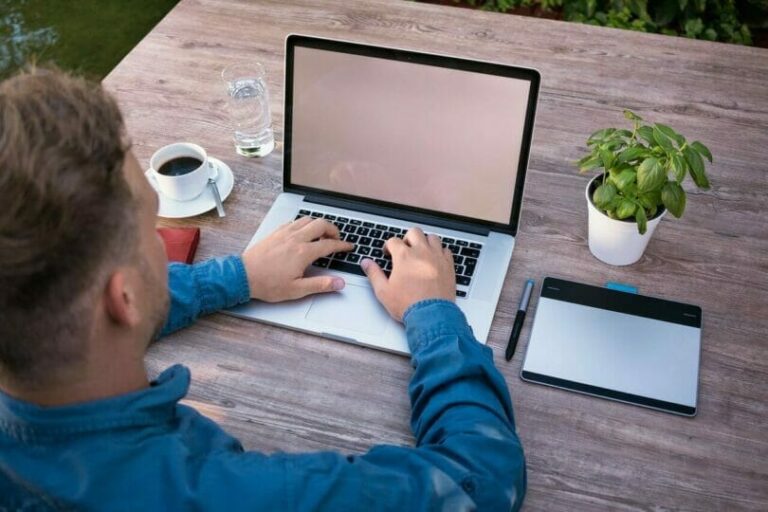 Person working on a laptop at a wooden table with a cup of coffee, a tablet, a pen, and a small plant.