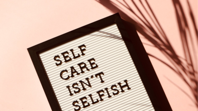 A board with "SELF CARE ISN'T SELFISH" written on it rests on a peach background with a shadow of palm leaves.