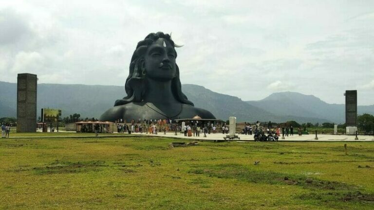 Large black statue of a meditating face with flowing hair set against a mountainous background, surrounded by visitors and greenery.