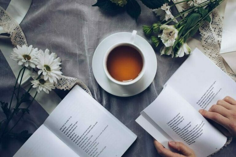 A cup of tea on a table surrounded by open books, white flowers, and lace cloth, with hands flipping the book pages.