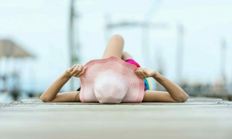 Person lying on a dock, wearing a pink sunhat and colorful swimsuit, holding the hat while facing the water, with legs slightly crossed.