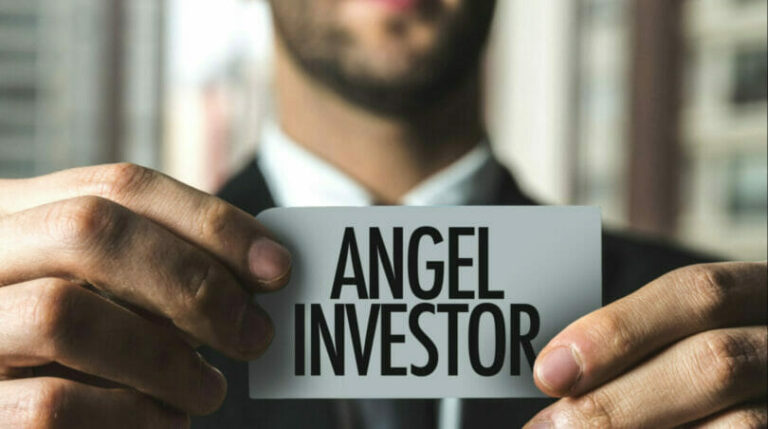 Man holding a business card that reads "ANGEL INVESTOR" in bold letters, with a blurred cityscape in the background.