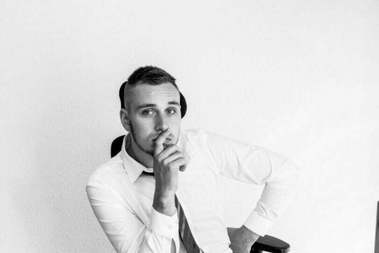 Man in a white shirt and tie sitting in a chair, leaning forward with hand on chin in a thoughtful pose.