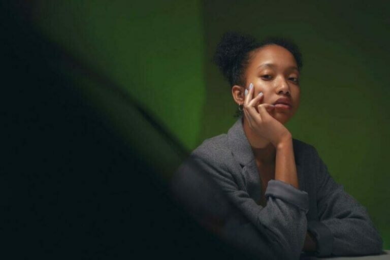 Woman in a gray blazer sits pensively with her chin resting on her hand against a green background.