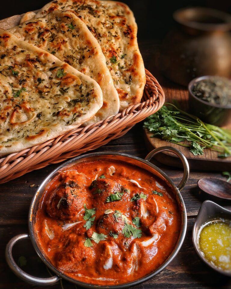 A bowl of creamy butter chicken garnished with cilantro, served with garlic naan bread in a basket, herbs, and butter on the side.