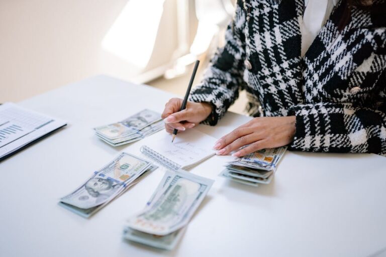 Person in plaid jacket writing on notepad at a desk, surrounded by stacks of US dollar bills and financial documents.