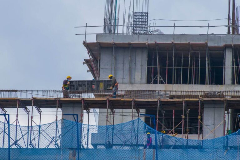 Construction workers in safety gear are working on a multi-story concrete building framed with scaffolding and blue safety nets.