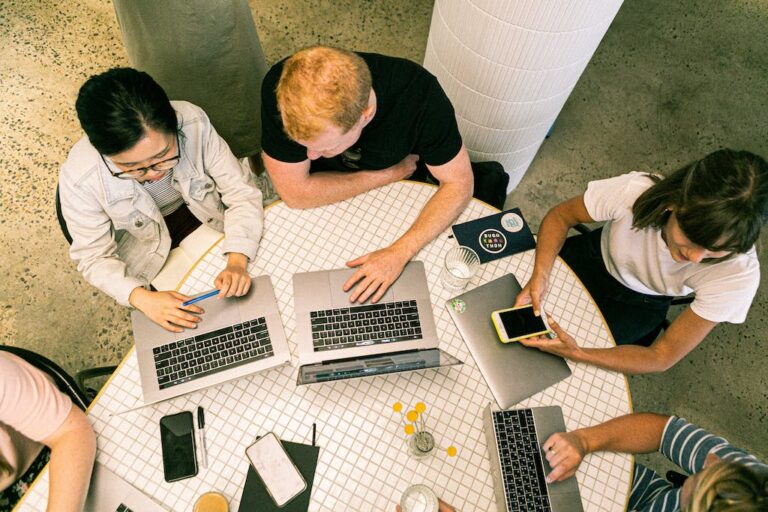 Four people collaborate around a table with laptops, phones, and notebooks. Top-down view in a modern workspace setting.