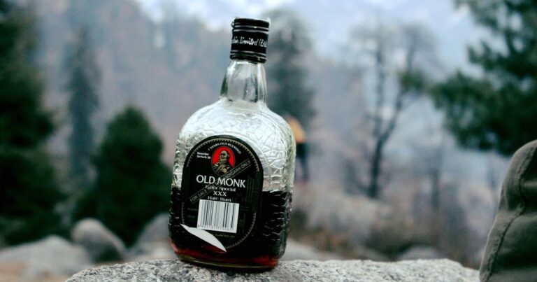 Old Monk Rum bottle on a rock, with blurred forest background.